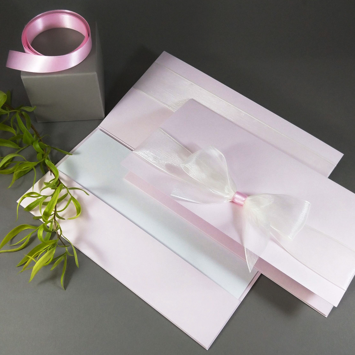 Dárková kapsa DL formátu z matného, mírně strukturovaného papíru světle růžové barvy, s aplikací z široké monofilové stuhy, celkový rozměr 206 × 106 mm.