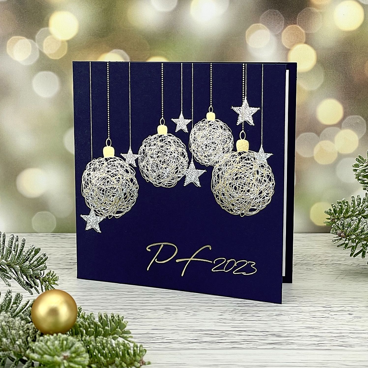 Novoroční–vánoční přání s motivem zavěšených zlatých a stříbrných baněk a hvězd, z tmavě modrého matného papíru, otevírací, čtvercový formát 135 × 135 mm.
