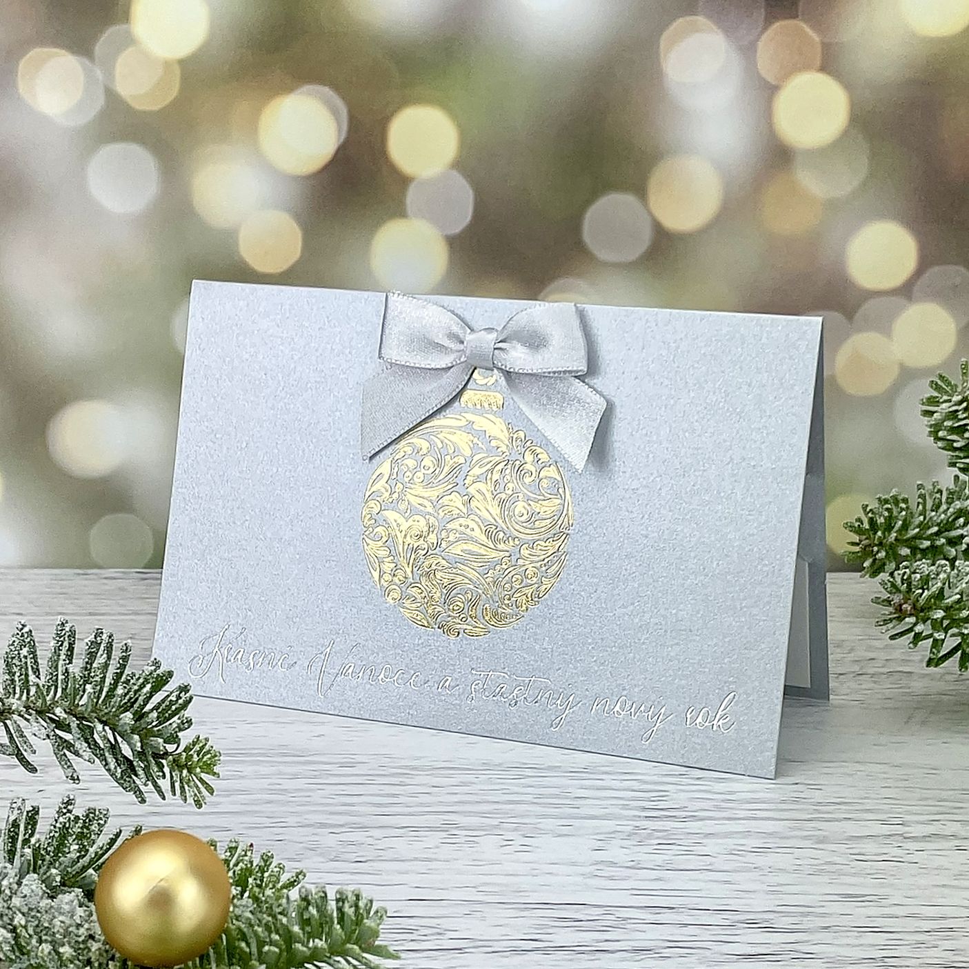Novoroční–vánoční přání s motivem zlaté reliéfní baňky s atlasovou mašlí, stříbrný metalický papír, otevírací, formát 155 × 100 mm.
