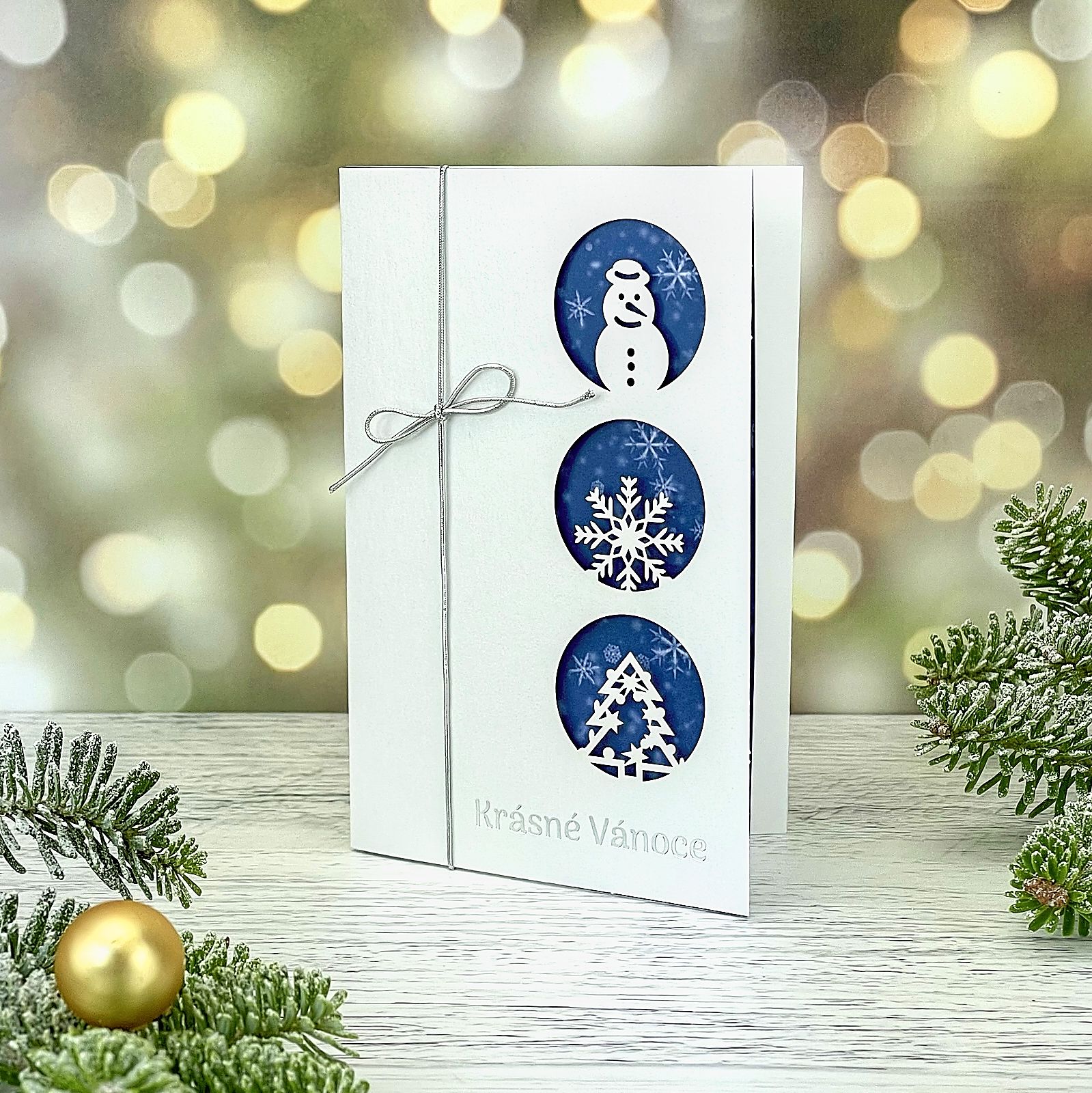 Novoroční–vánoční přání z bílého perleťového papíru s laserovanými zimními symboly - sněhulák, vločka, stromeček, doplněnými stříbrnou ražbou a mašličkou ze stříbrné pruženky; otevírací, formát 100 × 150 mm.