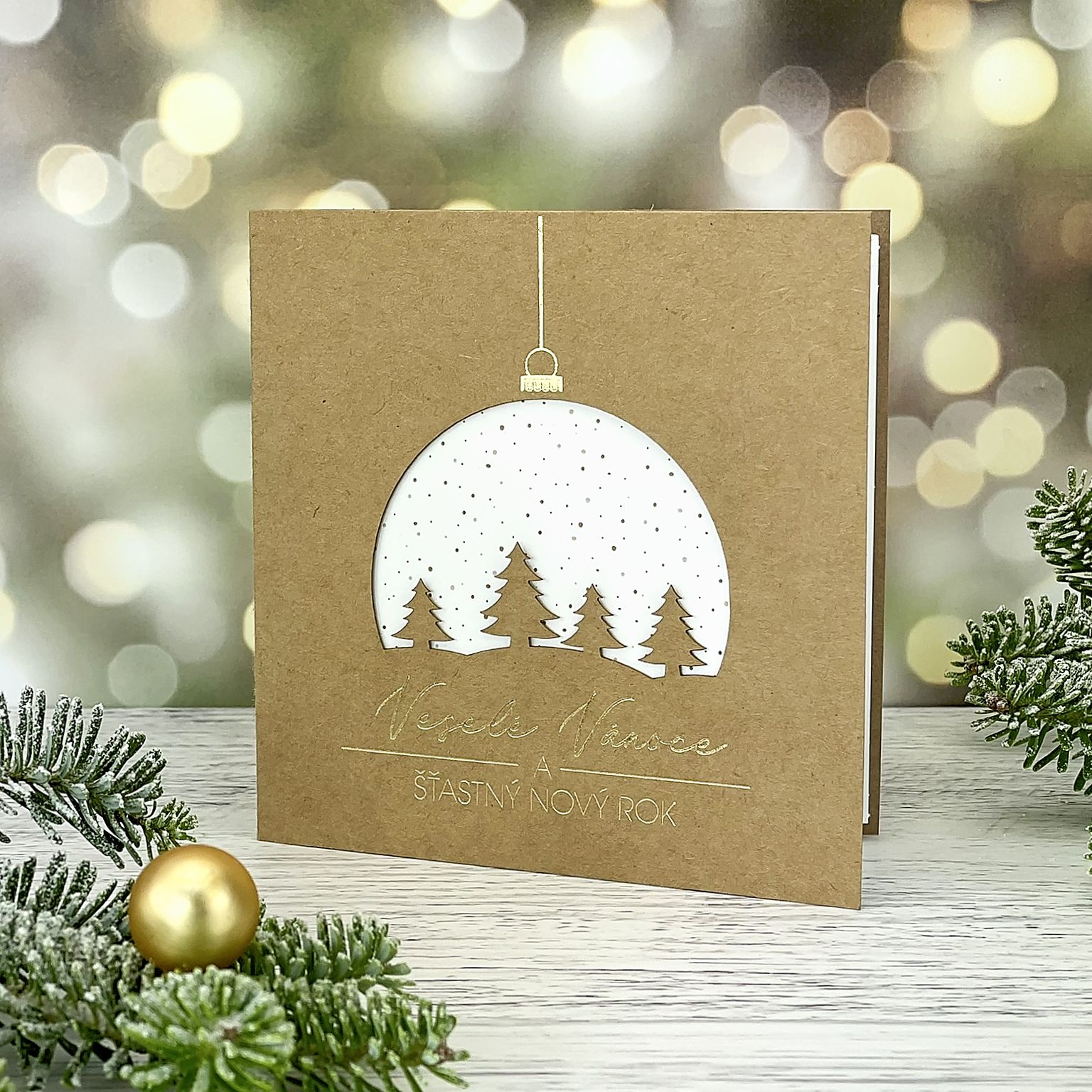 Novoroční–vánoční přání z recyklovaného papíru s laserovaným motivem zasněžené krajinky v baňce, otevírací, čtvercový formát 135 × 135 mm.