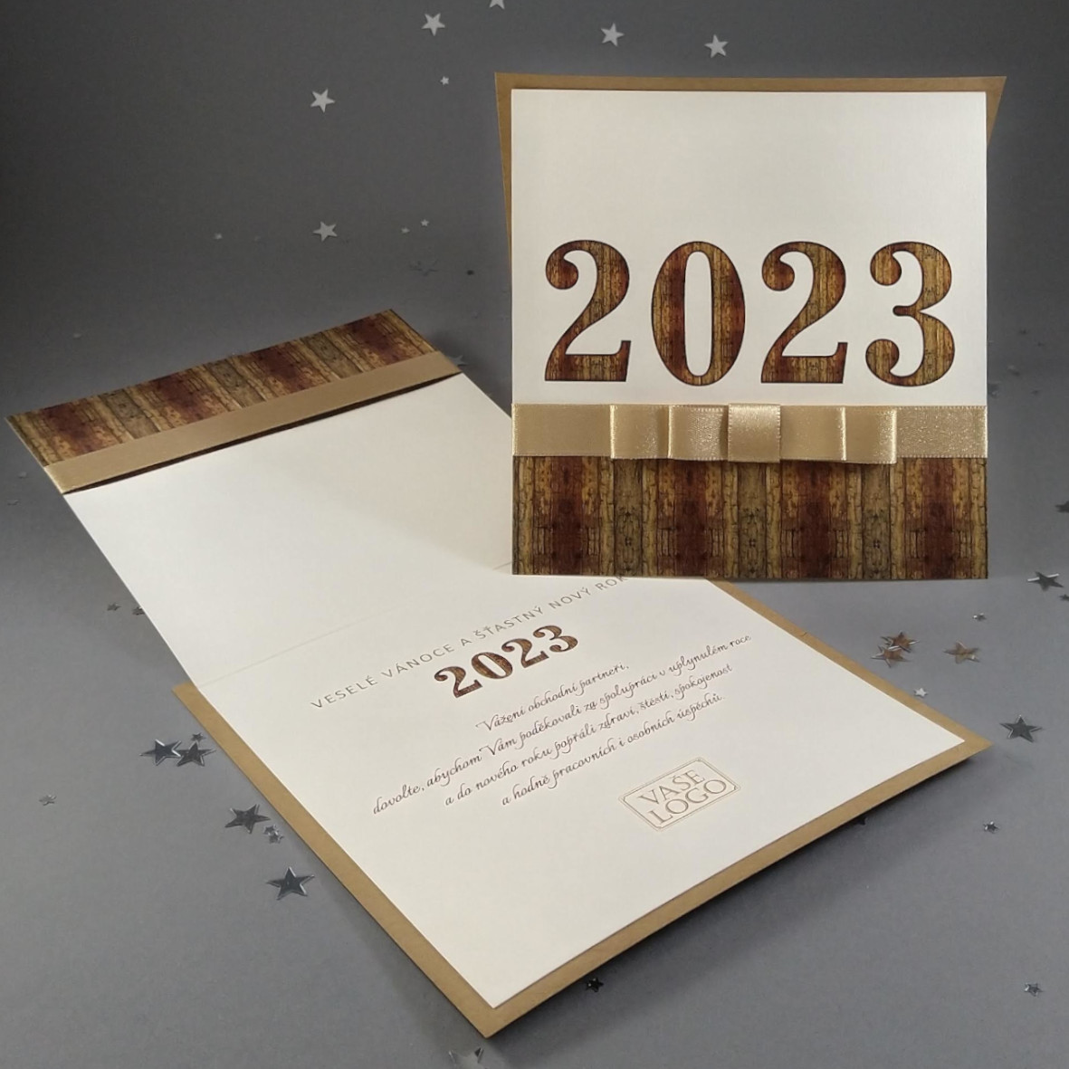 Novoroční–vánoční přání s motivem dřvěného letopočtu s atlasovou stuhou, čtvercový formát 146 × 146 mm, digitální tisk, kombinace recyklovaného a světle zlatého perleťového papíru.